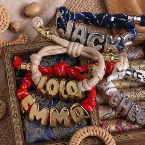 Op de afbeelding: Vier verstelbare koordarmbanden met metalen letterkralen die de namen JACKS, LOLA, EMMA en CHLOE spellen. De armbanden zijn gemaakt van marineblauw, beige, rood en wit patroonkoord met goud- en zilverkleurige hardware-accenten.