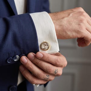 Op de afbeelding: Een ronde goudkleurige manchetknop met een kleine, zwart-wit vintage-stijl foto van twee kinderen. De manchetknop zit vast door de witte manchet van een overhemd met lange mouwen onder een donkerblauwe blazer met bijpassende knopen.