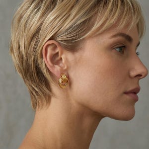 Peut inclure: Boucles d'oreilles dorées à clous avec un design de liens. Les boucles d'oreilles présentent trois cercles imbriqués, créant un aspect texturé et dimensionnel. Les boucles d'oreilles sont de couleur or vif et brillant.