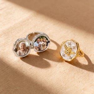 May include: Two rings on a beige surface. One is silver, with a hinged locket revealing a photo of a family and an elderly woman. The other is gold, featuring a cherub design with white enamel and small crystals.