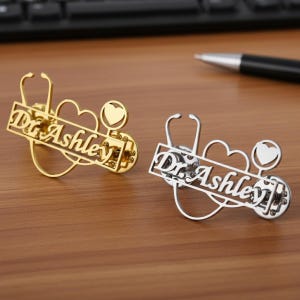 May include: Two metal pins, one gold and one silver, featuring a stethoscope, heart, and the text "Dr. Ashley". The pins are designed with a cutout style, and are likely intended for medical professionals.
