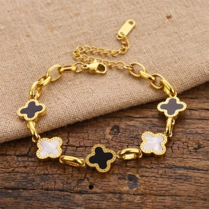 May include: Gold-coloured bracelet with a clover design. The bracelet has a chain-link style with alternating black and white clover-shaped charms. The bracelet is displayed on a textured, neutral-coloured surface.