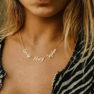 Op de afbeelding: Een goudkleurige ketting met de namen "Emily", "Mary" en "Sophia". De namen zijn in een cursief lettertype en verbonden door een fijne ketting. De ketting wordt gedragen tegen de huid van een persoon, met een zwart-wit patroon top zichtbaar.