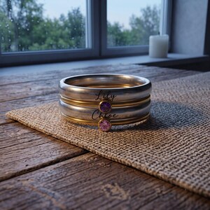 Pode incluir: Uma pilha de quatro anéis metálicos com bandas alternadas de prata e ouro. Duas bandas de prata estão gravadas com os nomes Lily e Chloe em letra cursiva preta. Duas bandas de ouro possuem cada uma uma pedra preciosa redonda, uma roxa e uma rosa.