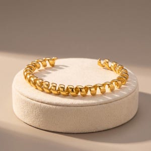 Op de afbeelding: Een goudkleurige armband met een doorlopend spiraal- of spiraalontwerp. Het metaal heeft een gepolijst, reflecterend oppervlak dat het licht opvangt en een reeks uniforme, ronde lussen vormt die de cirkelvorm van de open armband creëren.