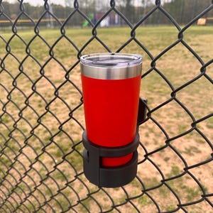 May include: A red insulated tumbler with a stainless steel lid is held in a black plastic cup holder attached to a chain link fence.