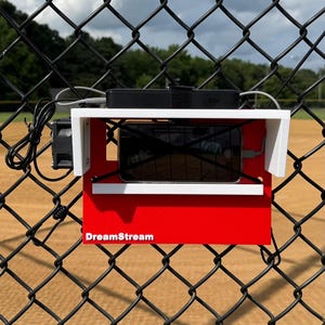 May include: A red and white device with a black screen is mounted on a chain-link fence. The device has the text "DreamStream" on the front. A black power bank and a small fan are attached to the device.