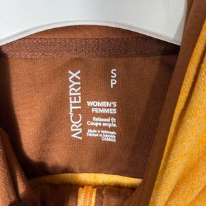 May include: Close-up of a brown and yellow hooded sweatshirt. The label reads "ARC'TERYX" and "WOMEN'S FEMMES". The garment has a relaxed fit and is made in Indonesia. The zipper is visible.