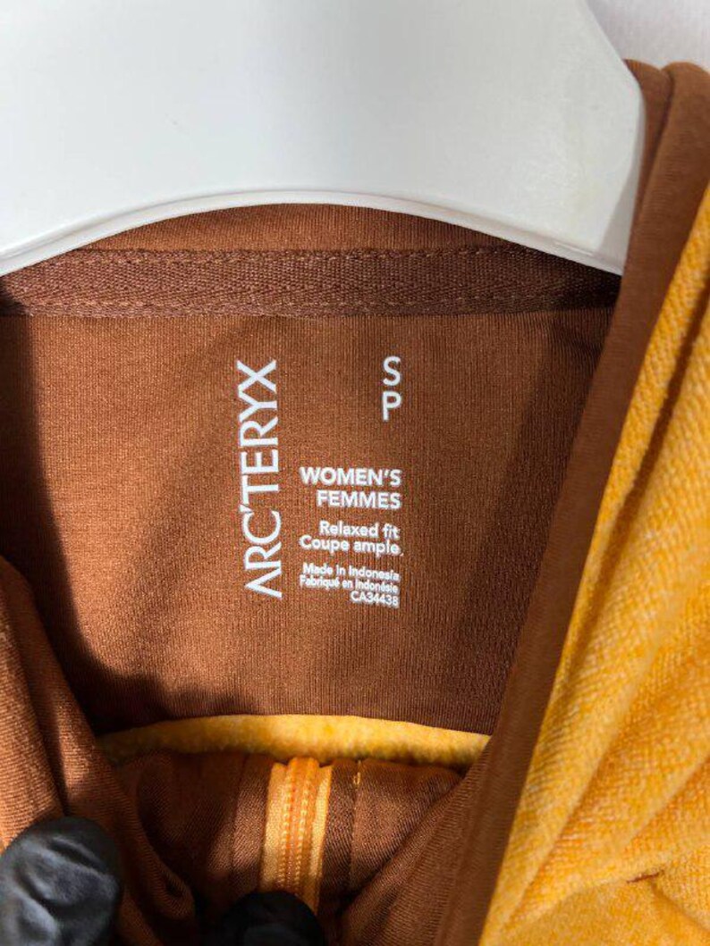 May include: Close-up of a brown and yellow hooded sweatshirt. The label reads "ARC'TERYX" and "WOMEN'S FEMMES". The garment has a relaxed fit and is made in Indonesia. The zipper is visible.