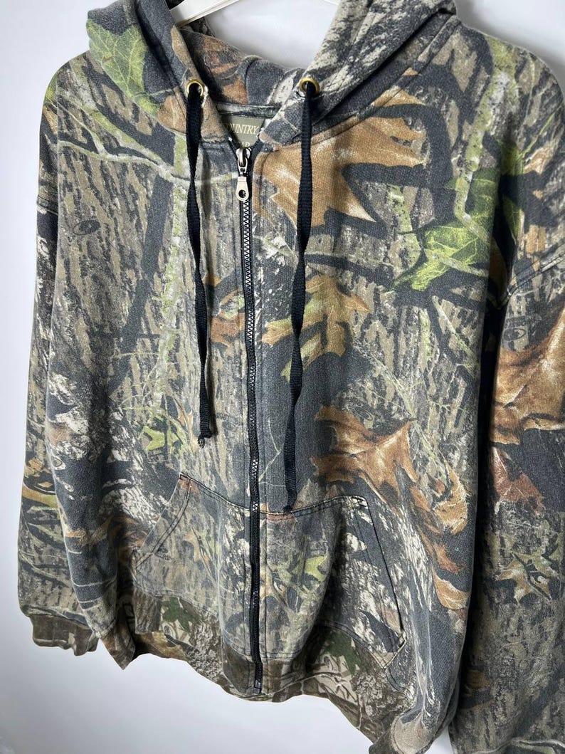Puede incluir: Sudadera con capucha y cremallera con estampado de camuflaje. La chaqueta tiene una capucha con cordones, una cremallera frontal completa y dos bolsillos delanteros. El dise&ntilde;o de camuflaje incorpora tonos de verde, marr&oacute;n y gris.