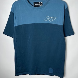 May include: Two-tone blue t-shirt with short sleeves. The top section is a lighter blue, while the bottom is a darker shade. Features the text "FP" in white script on the chest and the brand name "RAF SIMONS" near the neckline.