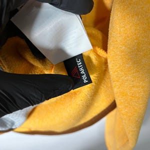 May include: Close-up of a bright yellow fleece garment. A black tag with the word "POLARTEC" in white is visible. The garment is held by a gloved hand, with a white tag partially visible.