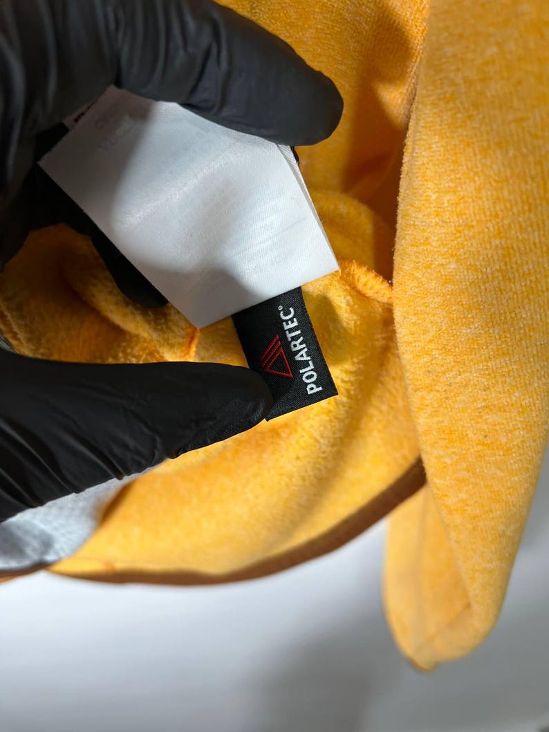 May include: Close-up of a bright yellow fleece garment. A black tag with the word "POLARTEC" in white is visible. The garment is held by a gloved hand, with a white tag partially visible.