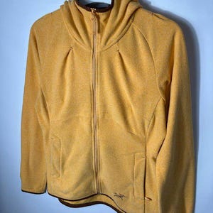 May include: Mustard yellow fleece jacket with a hood and a full-length zipper. The jacket has two front pockets and a brown trim around the hood and bottom hem. The jacket is hanging on a white hanger.