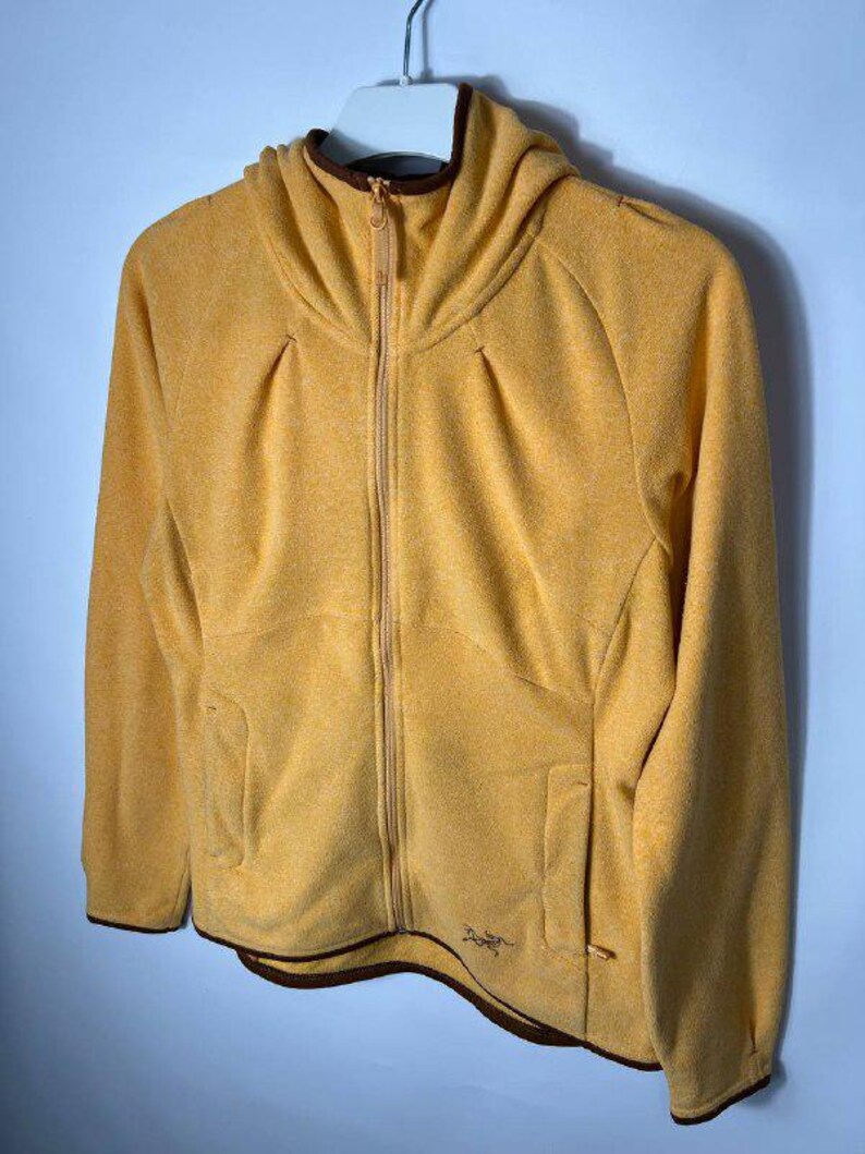 May include: Mustard yellow fleece jacket with a hood and a full-length zipper. The jacket has two front pockets and a brown trim around the hood and bottom hem. The jacket is hanging on a white hanger.