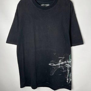 May include: Black short-sleeved t-shirt with a crew neck. The shirt features a white and green abstract design on the lower right side. The text on the collar is illegible.