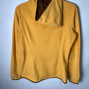 May include: Mustard yellow hooded sweatshirt with a brown interior lining and trim. The long-sleeved top is hanging on a white hanger against a light gray background. The sweatshirt has a textured appearance.