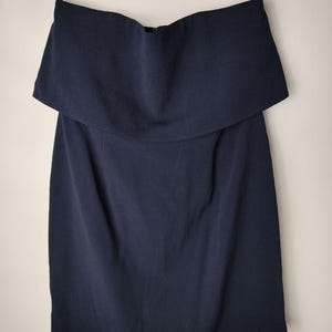 May include: A navy blue, strapless dress with a layered, off-the-shoulder neckline. The dress has a simple, elegant design and a knee-length hemline. The fabric appears to be a smooth, woven material. The dress is hanging from a black hanger.
