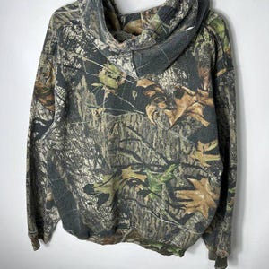 Puede incluir: Una sudadera con capucha con estampado de camuflaje y capucha con cord&oacute;n. Los colores incluyen tonos de marr&oacute;n, verde y beige, imitando un entorno forestal. La sudadera con capucha est&aacute; colgada de una percha blanca.