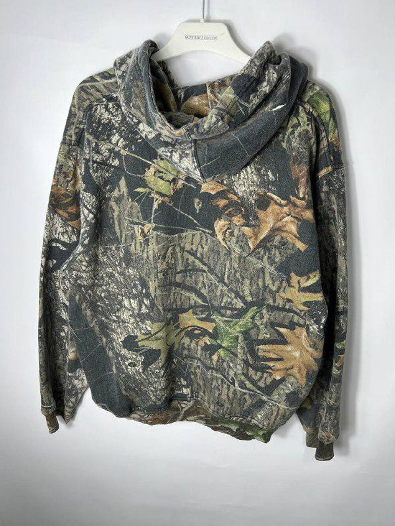 Puede incluir: Una sudadera con capucha con estampado de camuflaje y capucha con cord&oacute;n. Los colores incluyen tonos de marr&oacute;n, verde y beige, imitando un entorno forestal. La sudadera con capucha est&aacute; colgada de una percha blanca.