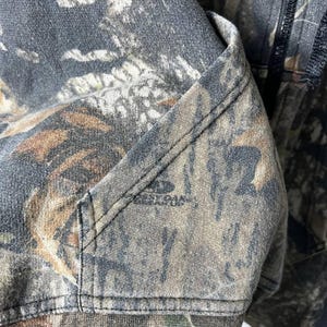 Puede incluir: Primer plano de una sudadera con capucha con estampado de camuflaje. La tela presenta una mezcla de verdes, marrones y grises, con un detalle de bolsillo visible y el nombre de la marca "BEST OAK" impreso en la tela.