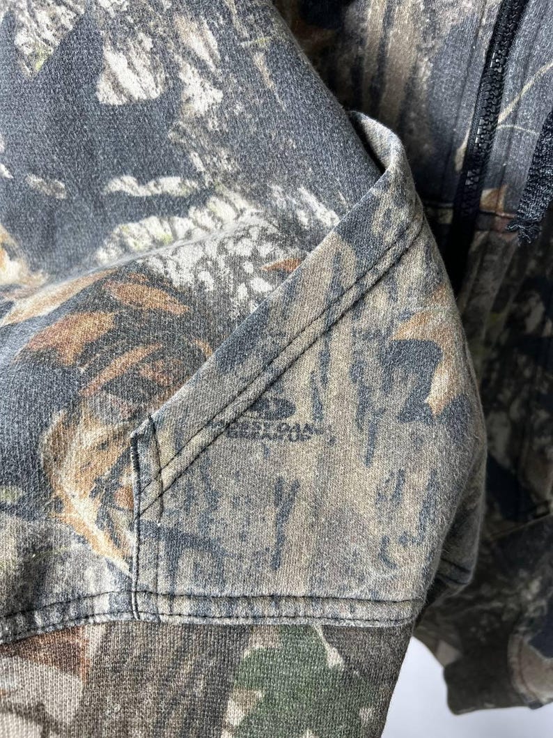 Puede incluir: Primer plano de una sudadera con capucha con estampado de camuflaje. La tela presenta una mezcla de verdes, marrones y grises, con un detalle de bolsillo visible y el nombre de la marca "BEST OAK" impreso en la tela.