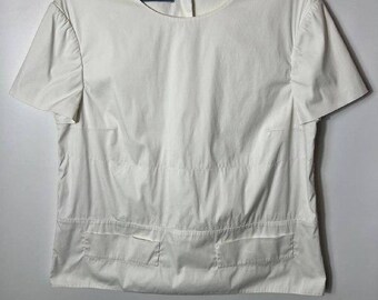 May include: A crisp white short-sleeved blouse featuring a round neckline and a horizontal seam across the midsection. The garment includes two rectangular flap pockets at the lower front and a Prada brand label visible at the inner collar.