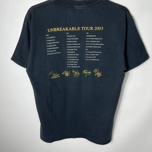 May include: A black short-sleeve t-shirt featuring yellow text that reads UNBREAKABLE TOUR 2003, followed by a list of concert dates and locations from April to July, and five gold-colored signatures printed across the bottom of the back.