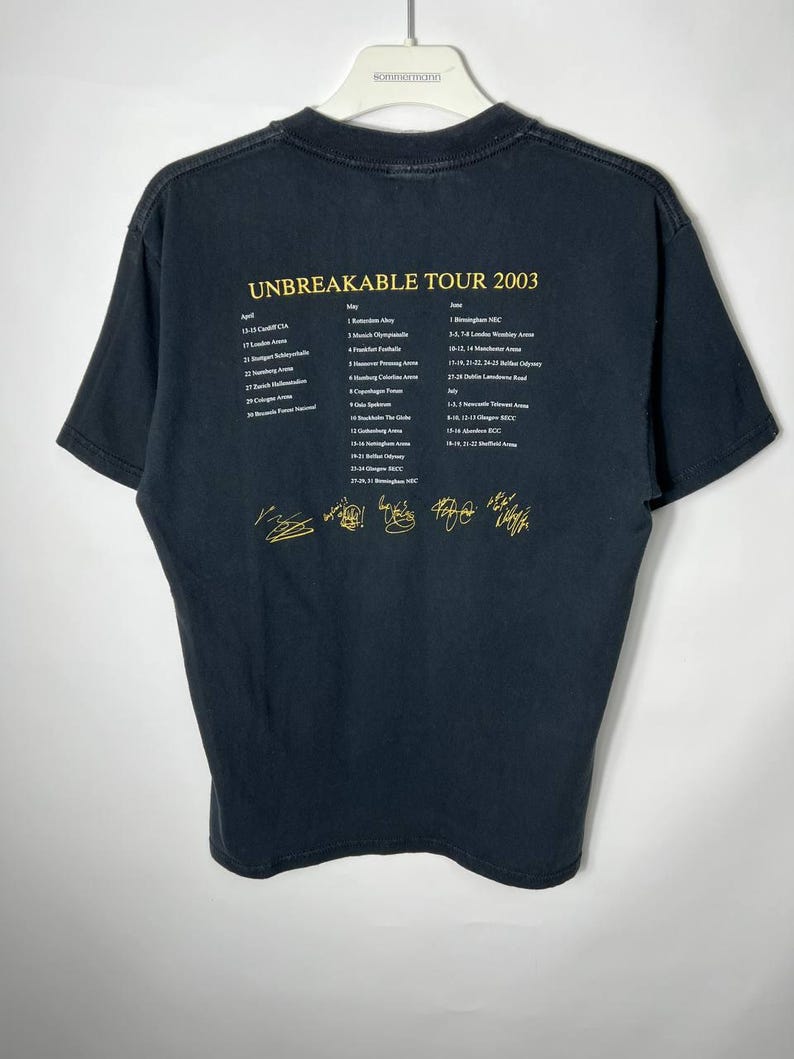 May include: A black short-sleeve t-shirt featuring yellow text that reads UNBREAKABLE TOUR 2003, followed by a list of concert dates and locations from April to July, and five gold-colored signatures printed across the bottom of the back.