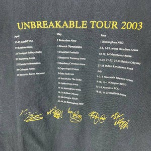 May include: A black t-shirt featuring yellow text that reads UNBREAKABLE TOUR 2003. Below the title, a list of concert dates and locations for April, May, June, and July is printed in white. Six yellow signatures are displayed at the bottom.