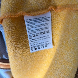 May include: Close-up of a yellow fabric with a tag attached. The tag contains text in multiple languages, including English, French, Spanish, and German, detailing the fabric's composition: 100% polyester and 79% polyester/21% elastane.