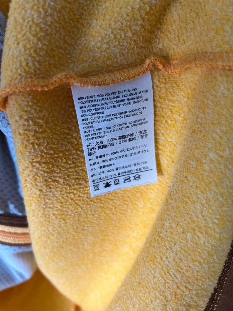 May include: Close-up of a yellow fabric with a tag attached. The tag contains text in multiple languages, including English, French, Spanish, and German, detailing the fabric's composition: 100% polyester and 79% polyester/21% elastane.