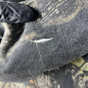 Puede incluir: Primer plano de una tela con estampado de camuflaje, principalmente en tonos grises, marrones y verdes. La tela parece ser un material texturizado, posiblemente forro polar o un textil similar. Se puede ver un peque&ntilde;o desgarro en la superficie.