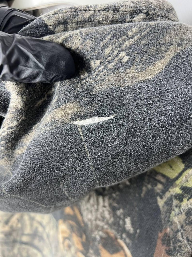 Puede incluir: Primer plano de una tela con estampado de camuflaje, principalmente en tonos grises, marrones y verdes. La tela parece ser un material texturizado, posiblemente forro polar o un textil similar. Se puede ver un peque&ntilde;o desgarro en la superficie.