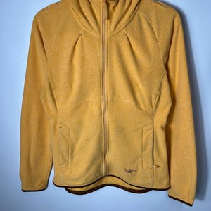 May include: Mustard yellow zip-up hooded jacket with a brown trim. The jacket features a full-length zipper, two front pockets, and a small logo on the lower right. The hood has a structured design. The jacket is made of a textured material.
