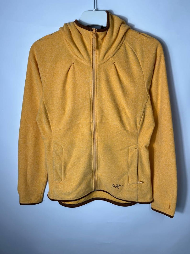 May include: Mustard yellow zip-up hooded jacket with a brown trim. The jacket features a full-length zipper, two front pockets, and a small logo on the lower right. The hood has a structured design. The jacket is made of a textured material.