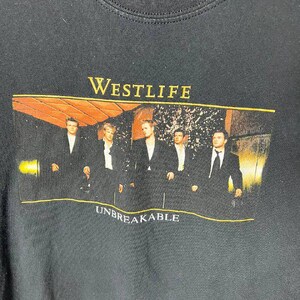 May include: A black crew neck t-shirt featuring a rectangular graphic print of the band Westlife. The print shows five individuals in black suits with the yellow text WESTLIFE above and the white text UNBREAKABLE below the group image.