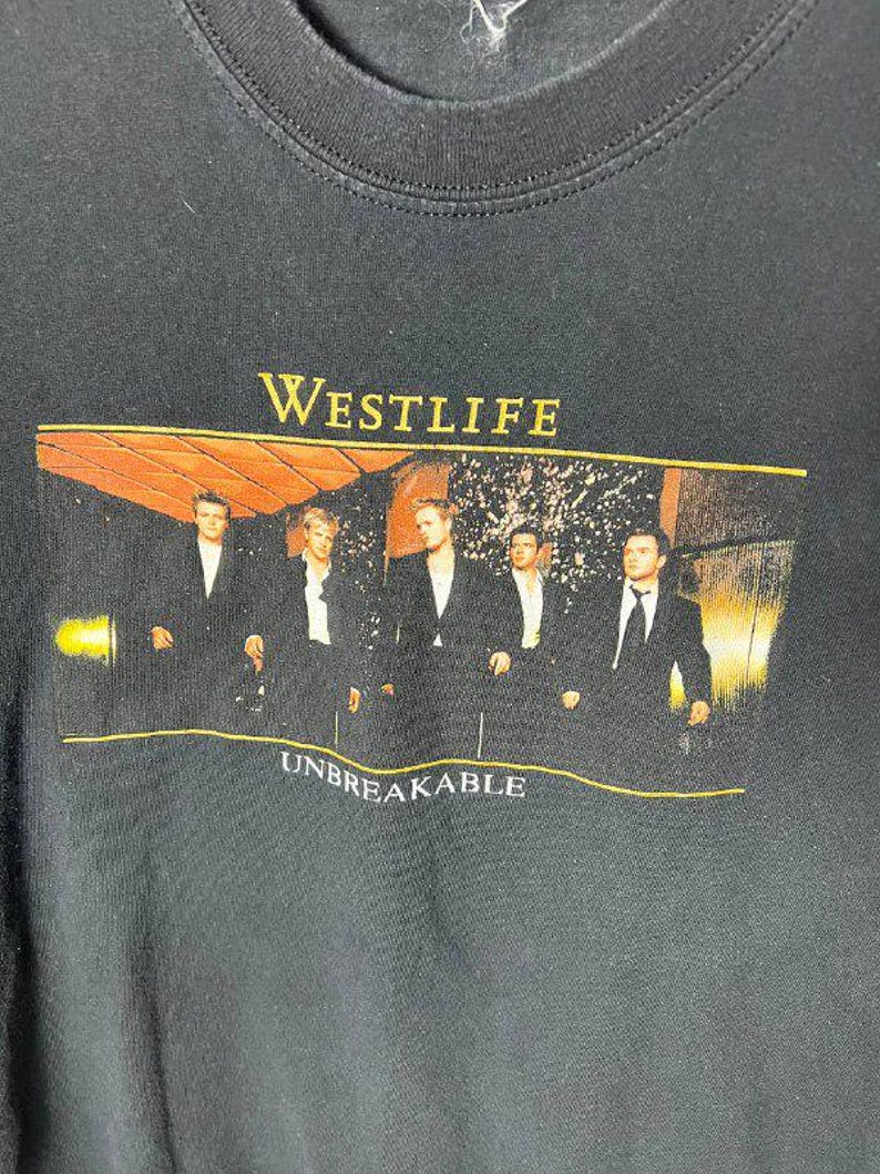 May include: A black crew neck t-shirt featuring a rectangular graphic print of the band Westlife. The print shows five individuals in black suits with the yellow text WESTLIFE above and the white text UNBREAKABLE below the group image.