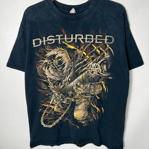 May include: A black short-sleeve t-shirt featuring a graphic print of a hooded, skeletal figure playing a guitar. The word DISTURBED is printed in white lettering above the figure, which is surrounded by yellow and orange flame-like accents and chains.