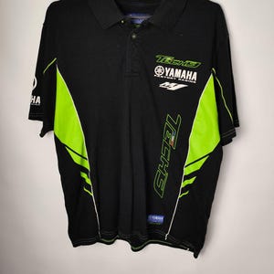 Peut inclure: Polo noir avec des accents vert fluo et le texte "TECH5 YAMAHA FACTORY RACING". Le t-shirt a un col et des manches courtes. Le logo Yamaha est visible sur la poitrine.