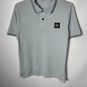 Può includere: Una polo a maniche corte grigio chiaro con una toppa a bussola Stone Island in bianco e nero sul petto sinistro. Il capo presenta un'abbottonatura a due bottoni, un colletto con bordini neri e strisce nere abbinate sui polsini delle maniche.