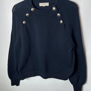 May include: A navy blue long-sleeve knit sweater featuring a ribbed crew neckline, ribbed cuffs, and a ribbed hem. The garment is accented with four polished silver-tone metal buttons arranged in a vertical row along the upper right shoulder area.
