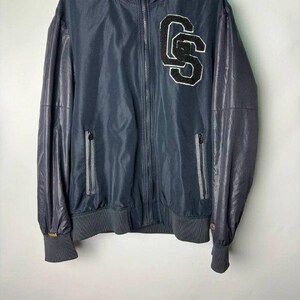 May include: Navy blue zip-up jacket with a high collar and ribbed cuffs. The jacket features a large "CS" patch on the chest and contrasting dark grey sleeves. Two zippered pockets are visible on the front.