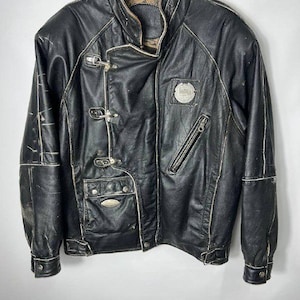 May include: A black leather jacket featuring a high collar, three metal toggle closures, a diagonal zipper pocket on the chest, and a lower flap pocket. The jacket includes white contrast stitching along the seams and two circular metal emblems.
