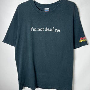 May include: Dark gray t-shirt with the text "I'm not dead yet" in white. The shirt has short sleeves and a crew neck. A small graphic is visible on the right sleeve.