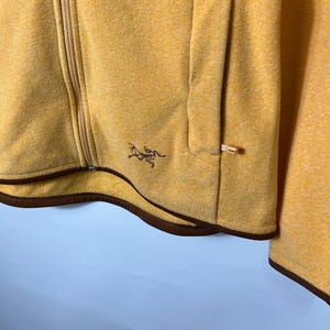 May include: Mustard yellow zip-up jacket with a brown trim and a small logo on the chest. The jacket has a pocket on the front and a full-length zipper. The fabric appears to be a soft, textured material.