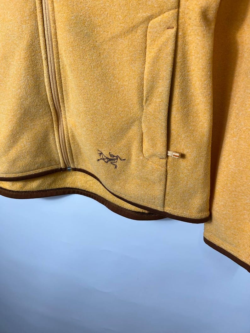 May include: Mustard yellow zip-up jacket with a brown trim and a small logo on the chest. The jacket has a pocket on the front and a full-length zipper. The fabric appears to be a soft, textured material.