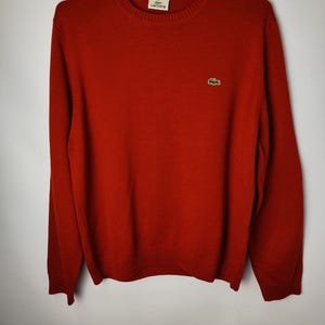 May include: A red crew neck sweater with long sleeves. The sweater has a ribbed hem and cuffs. A small green crocodile logo is on the left chest. The brand name "Lacoste" is on a tag at the neckline.