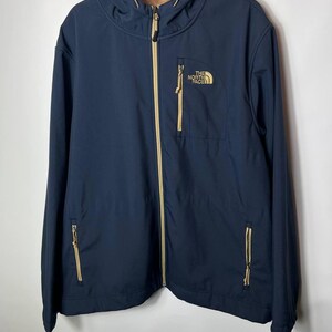May include: A navy blue hooded jacket featuring a tan front zipper, tan zipper pulls, and two lower zippered pockets. A tan zippered chest pocket displays the white text The North Face logo. The interior is lined with a tan fleece material.