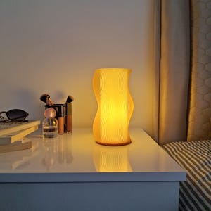 Könnte beinhalten: Eine wellenförmige, gelbe Nachttischlampe beleuchtet einen weißen Nachttisch. Die Lampe hat ein vertikales, geripptes Design. Weitere Gegenstände auf dem Nachttisch sind Make-up-Pinsel, Parfüm und Sonnenbrillen. Die Lampe ist etwa 25 cm hoch.